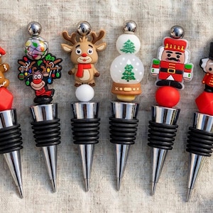 Könnte beinhalten: Ein Set aus sechs Flaschenverschlüssen aus Metall mit schwarzen Gummiringen, jeweils gekrönt von einer festlichen Silikonfigur, darunter ein Lebkuchenmann, ein Rentier, eine Schneekugel mit Bäumen und zwei Nussknacker-Figuren.