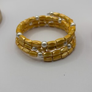 May include: A spiral bracelet with three strands of yellow, geometric beads. The bracelet is accented with small, round, white beads. The jewelry is displayed on a white surface.