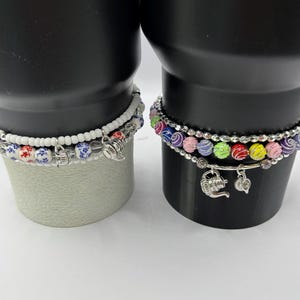 May include: Two beaded bracelets featuring silver-toned charms. The left bracelet has white beads and blue and red floral patterned beads. The right bracelet features multi-colored beads in purple, pink, green, red, and yellow with silver accents.
