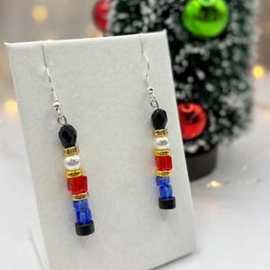 May include: A pair of dangle earrings featuring a vertical arrangement of beads. Each earring consists of a black bead, a gold-tone spacer, a white pearl-like bead, a gold-tone spacer, a red cube bead, a gold-tone spacer, and a blue cube bead.