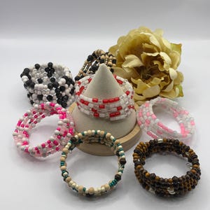 Pode incluir: Uma coleção de pulseiras de contas em várias cores e designs. As pulseiras incluem estilos de contas em preto e branco, vermelho e branco, rosa e branco e de madeira. Uma flor amarela está no fundo.