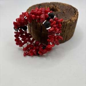 May include: A red coral bracelet with black lava beads and silver-toned details. The bracelet is composed of small, irregularly shaped coral pieces strung together. The lava beads are round and textured. The bracelet is displayed on a wooden surface.