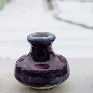 May include: A small, handcrafted ceramic vase with a bulbous body and a narrow neck. The vase is glazed in a deep purple color with blue accents around the rim and base. The vase is approximately 3 inches tall.