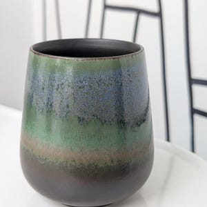 Può includere: Un vaso in ceramica con una sfumatura di colori, che passa dal marrone scuro alla base alle tonalità verdi e blu verso l'alto. L'interno è nero e il vaso ha una forma liscia e arrotondata.