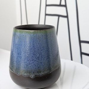Könnte beinhalten: Eine handgefertigte Keramikvase mit einer einzigartigen Form. Die Vase weist einen Farbverlauf auf, der von einem dunkelbraunen Sockel zu einem blau-grünen oberen Bereich übergeht. Der Innenraum ist dunkel. Die Vase wird wahrscheinlich für die Inneneinrichtung verwendet.