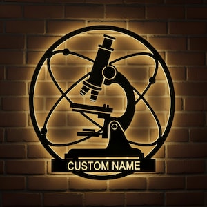Puede incluir: Arte de pared de metal negro con un diseño de microscopio dentro de una estructura atómica circular. Las palabras "CUSTOM NAME" se muestran en una placa rectangular debajo del microscopio. El arte está iluminado contra una pared de ladrillos.