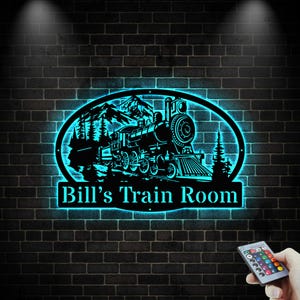 May include: A black metal train-themed wall art piece with a blue neon glow. The design features a train, mountains, and trees inside an oval frame. The text "Bill's Train Room" is below the frame.