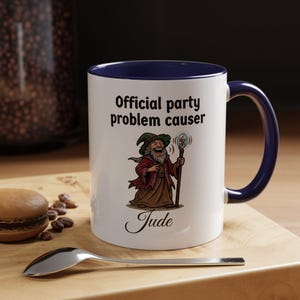 Peut inclure: Mug en céramique blanche avec intérieur et anse bleu marine. Le mug présente le texte "Official party problem causer" et une illustration de dessin animé de magicien. Le nom "Jude" est imprimé en dessous.