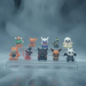 Può includere: Una collezione di piccole e colorate statuette di vari personaggi dei cartoni animati, tra cui un fantasma, un pipistrello e una strega. Le statuette sono disposte su un espositore trasparente a più livelli, su uno sfondo nebbioso.