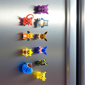 Könnte beinhalten: Eine Sammlung farbenfroher, cartoonartiger Magnete in verschiedenen Charakterdesigns, darunter ein lila katzenartiges Wesen, eine blau-weiße Figur und ein gelber mausartiger Charakter. Die Magnete sind an einer silbernen Kühlschranktür befestigt.