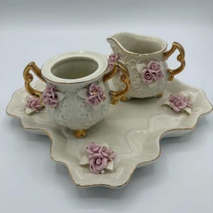 May include: A cream-colored porcelain tea set with gold accents and pink rose embellishments. The set includes a sugar bowl, creamer, and a decorative tray. The tray and the set are trimmed with gold.