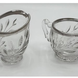 May include: Two-piece clear glass creamer and sugar bowl set. Both pieces have a silver rim and handles. The glass is etched with a leaf design. The creamer has a spout, and the sugar bowl has two handles.