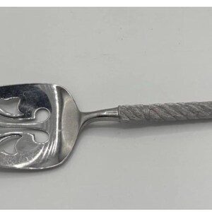 May include: A silver cake server with a decorative floral design on the blade. The handle has a textured, twisted rope design. The server is made of a shiny, reflective metal.