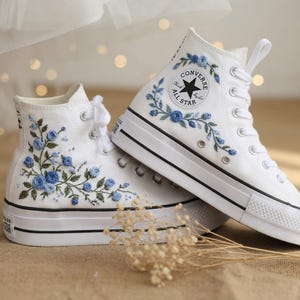 Peut inclure: Baskets montantes blanches avec broderie florale bleue. Les baskets présentent une semelle noire et blanche, des lacets blancs et le logo Converse All Star. Le motif floral comprend des roses bleues et des feuilles vertes, ajoutant une touche décorative.