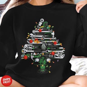 May include: Black sweatshirt featuring a Christmas tree design made of various tools, including wrenches, pliers, and sockets. The tree is decorated with red ornaments and string lights, with a green power tool at the base.