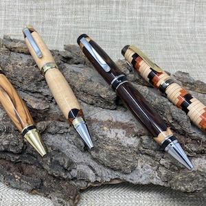 May include: Four handcrafted pens with distinct designs. The first is light brown with dark grain, the second is pale wood, the third is dark polished wood, and the fourth features a multi-colored geometric mosaic pattern. Each has a metal clip.