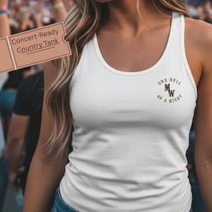 Puede incluir: Camiseta sin mangas blanca con el texto "ONE HELL OF A NIGHT" en letras marrones. La camiseta sin mangas es un tanque country listo para conciertos. La imagen está tomada en un concierto con una multitud al fondo.