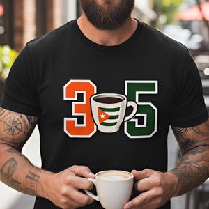 May include: Black t-shirt featuring the numbers "3" and "5" in orange and green, with a coffee cup graphic in between. The coffee cup has a Cuban flag design. The person in the image is holding a white coffee cup.