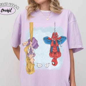 Op de afbeelding: Een lichtpaars Comfort Colors Orchid T-shirt met een cartoonillustratie van Rapunzel en Spider-Man die ondersteboven hangen. Rapunzel houdt haar lange blonde haar vast en Spider-Man hangt aan een web. Spraakballonnen met "O HAI!" en een vraagteken.