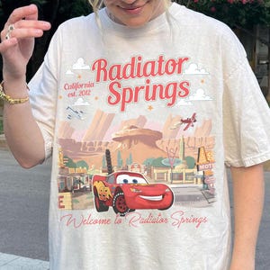 May include: Cream-colored t-shirt featuring a graphic of Radiator Springs with Lightning McQueen. The design includes the text "Radiator Springs" in red, "California est. 2012", and "Welcome to Radiator Springs".