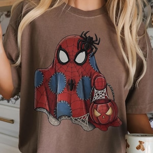 May include: Brown t-shirt featuring a Spiderman-themed ghost design with a spider on top. The ghost is red with a spiderweb pattern and blue patches. A pumpkin-shaped basket with a spiderweb design is also visible.