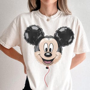 Puede incluir: Camiseta color crema con un dise&ntilde;o de globo de Mickey Mouse. El globo tiene orejas negras y una cara sonriente con una cuerda roja. La camiseta es de corte holgado.