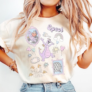 May include: Cream-colored t-shirt featuring a collection of whimsical illustrations. The design includes a purple dragon, the word "Epcot", a rainbow, and the phrase "one little spark". The shirt is paired with blue jeans.
