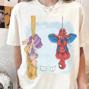 Op de afbeelding: Cr&egrave;mekleurig T-shirt met een cartoonillustratie van Rapunzel en Spider-Man die ondersteboven hangen. Rapunzel houdt haar lange blonde haar vast en Spider-Man hangt aan een web. Spraakballonnen zeggen "O HAI!" en "?".