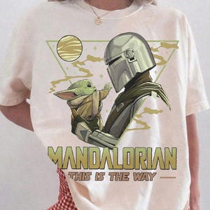 May include: Ivory-colored t-shirt featuring a graphic of The Mandalorian and Grogu. The design includes the text "MANDALORIAN" and "THIS IS THE WAY". The shirt is a Comfort Colors brand.