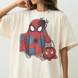 May include: Cream-colored t-shirt featuring a Halloween-themed graphic. The design depicts a ghost in a Spider-Man costume holding a pumpkin-shaped candy bucket. The ghost is red and blue with a spider web design.