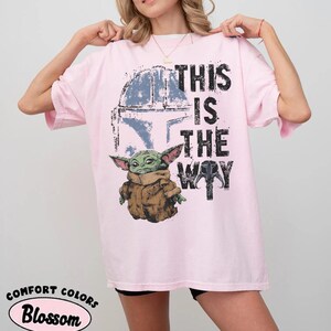 May include: Light pink t-shirt featuring a graphic of a green alien character in a brown coat, with the text "THIS IS THE WAY" in black. The design includes a blue helmet. The shirt is from Comfort Colors.