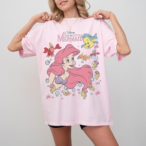 Pode incluir: Camiseta rosa claro com um desenho de Ariel de A Pequena Sereia da Disney. O design inclui Ariel com cabelo rosa, um suti&atilde; de concha roxa e v&aacute;rias criaturas marinhas. A camiseta tem mangas curtas e gola redonda.