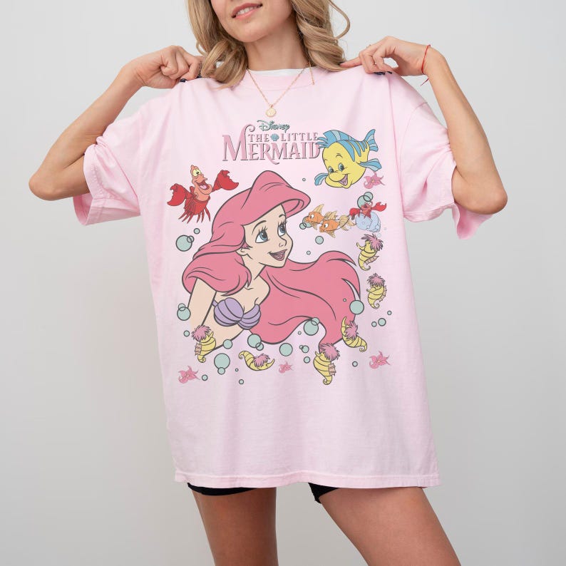 Pode incluir: Camiseta rosa claro com um desenho de Ariel de A Pequena Sereia da Disney. O design inclui Ariel com cabelo rosa, um suti&atilde; de concha roxa e v&aacute;rias criaturas marinhas. A camiseta tem mangas curtas e gola redonda.