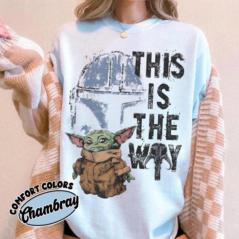 May include: Light blue sweatshirt with a graphic of a green alien character and a helmet, with the text "THIS IS THE WAY". The sweatshirt has the words "COMFORT COLORS" and "Chambray" in a circular design.
