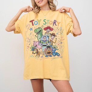 May include: Yellow t-shirt featuring the words "Toy Story" in colorful letters above a graphic of characters from the animated film. The design includes Woody, Buzz Lightyear, and other beloved characters. The shirt has a relaxed fit.