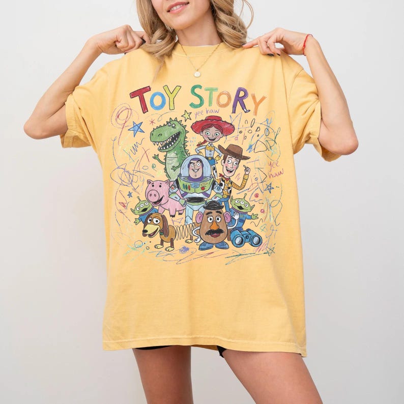 May include: Yellow t-shirt featuring the words "Toy Story" in colorful letters above a graphic of characters from the animated film. The design includes Woody, Buzz Lightyear, and other beloved characters. The shirt has a relaxed fit.