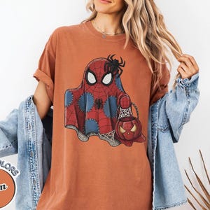 May include: Orange Comfort Colors t-shirt featuring a Spiderman ghost design with a spider and a pumpkin-shaped candy bucket. The shirt has a round neck and short sleeves. The shirt is part of the Yam Comfort Colors collection.
