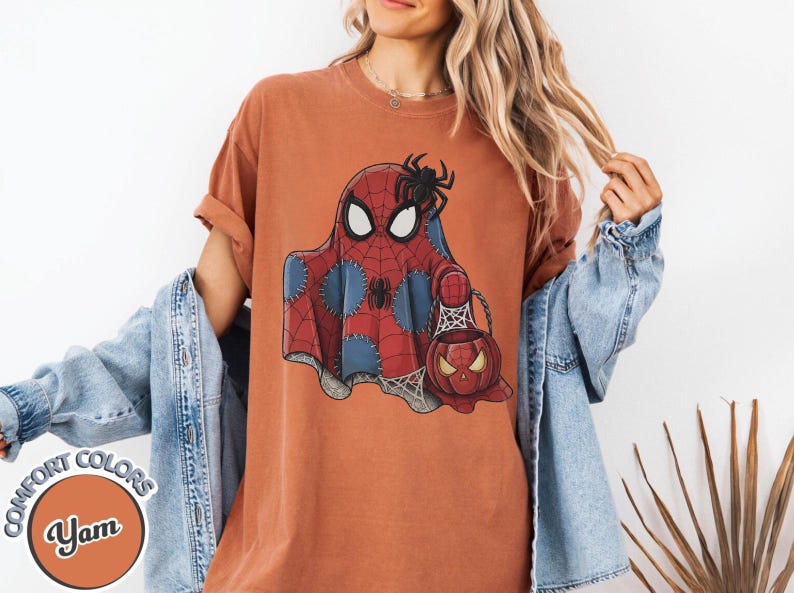 May include: Orange Comfort Colors t-shirt featuring a Spiderman ghost design with a spider and a pumpkin-shaped candy bucket. The shirt has a round neck and short sleeves. The shirt is part of the Yam Comfort Colors collection.