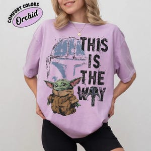 May include: Orchid-colored t-shirt featuring a graphic of Grogu, a Mandalorian helmet, and the phrase "THIS IS THE WAY" in black. The shirt has a crew neck and short sleeves. The text "COMFORT COLORS" and "Orchid" are in a pink oval.