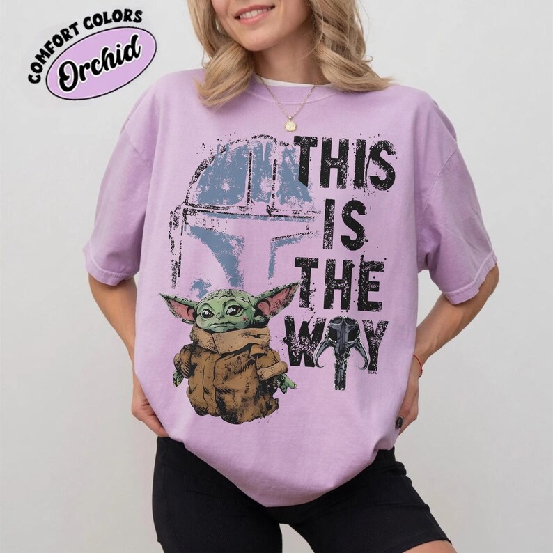 May include: Orchid-colored t-shirt featuring a graphic of Grogu, a Mandalorian helmet, and the phrase "THIS IS THE WAY" in black. The shirt has a crew neck and short sleeves. The text "COMFORT COLORS" and "Orchid" are in a pink oval.