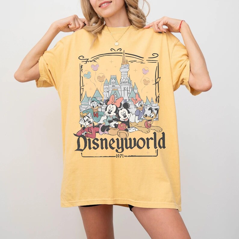May include: Yellow t-shirt featuring a vintage Disney World graphic. The design includes Mickey and Minnie Mouse, Donald Duck, and other characters in front of a castle. The text "Disneyworld 1971" is also visible.