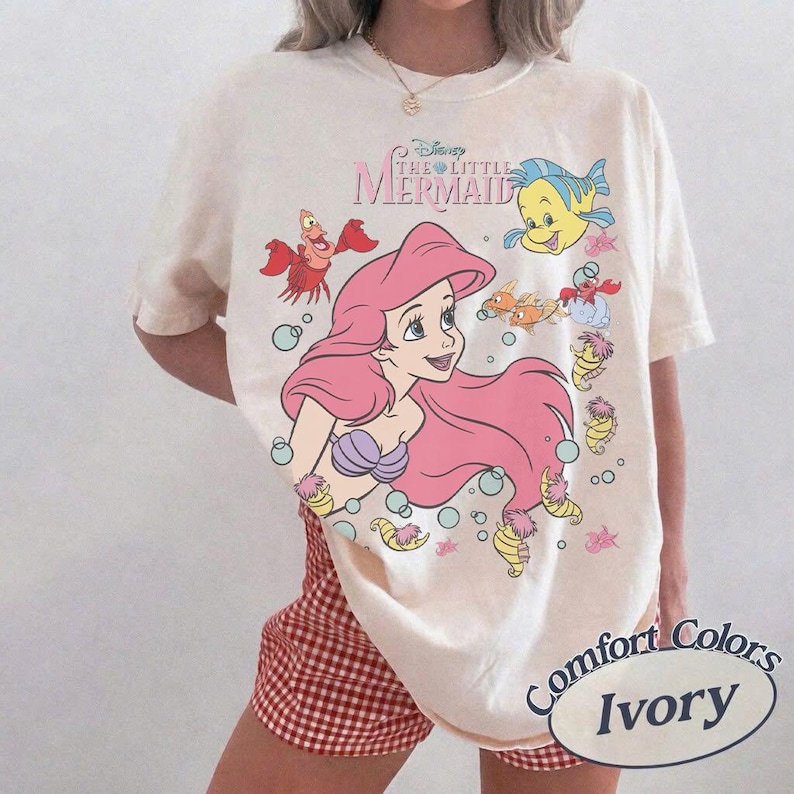 Pode incluir: Camiseta cor marfim com um gr&aacute;fico de Ariel da Pequena Sereia da Disney. O design inclui Ariel com cabelo rosa, Linguado, Sebasti&atilde;o e outras criaturas marinhas. A camisa &eacute; rotulada como "Comfort Colors" e "Ivory".