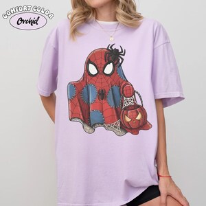 May include: A light purple Comfort Color t-shirt featuring a Halloween-themed Spider-Man ghost design. The graphic includes a red and blue ghost with a spider on top, holding a jack-o'-lantern bag. The shirt has the word "Orchid" printed on it.