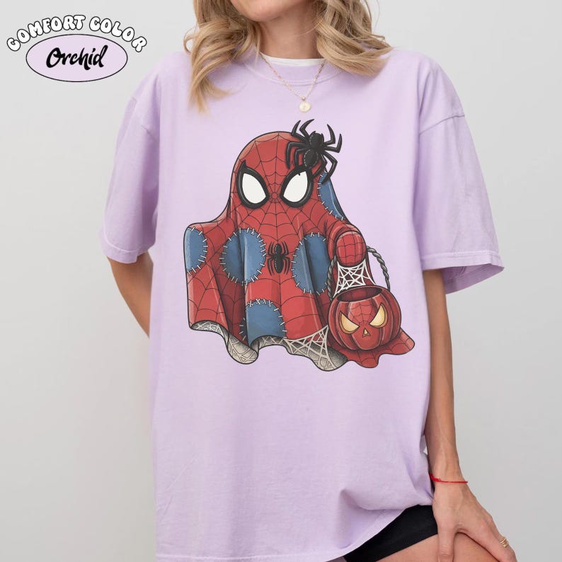 May include: A light purple Comfort Color t-shirt featuring a Halloween-themed Spider-Man ghost design. The graphic includes a red and blue ghost with a spider on top, holding a jack-o'-lantern bag. The shirt has the word "Orchid" printed on it.