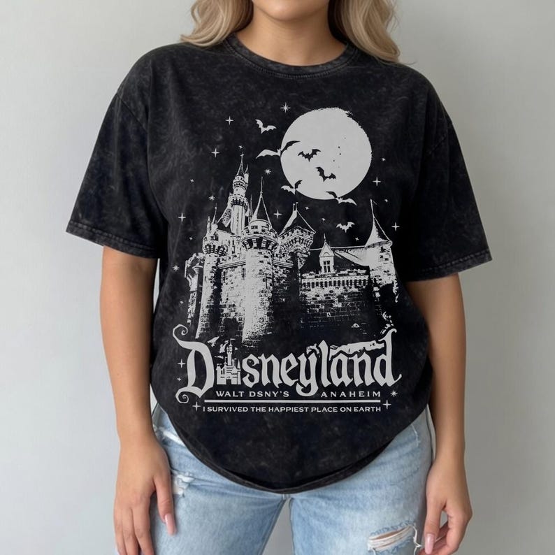 Pode incluir: Camiseta preta com um gr&aacute;fico branco de um castelo, lua e morcegos. A palavra "Disneyland" &eacute; exibida em uma fonte estilizada, com "Walt Dsny's Anaheim" e "I survived the happiest place on earth" abaixo.