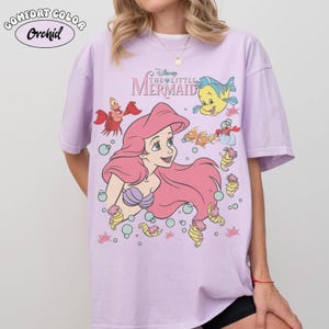 Pode incluir: Uma camiseta lil&aacute;s Comfort Colors com Ariel de A Pequena Sereia da Disney. O gr&aacute;fico inclui Ariel com cabelo rosa, um suti&atilde; de concha roxa e v&aacute;rias criaturas marinhas. A camisa tem o texto "The Little Mermaid".