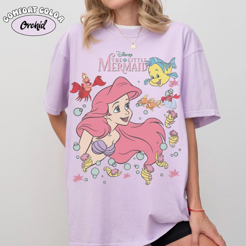 Pode incluir: Uma camiseta lil&aacute;s Comfort Colors com Ariel de A Pequena Sereia da Disney. O gr&aacute;fico inclui Ariel com cabelo rosa, um suti&atilde; de concha roxa e v&aacute;rias criaturas marinhas. A camisa tem o texto "The Little Mermaid".
