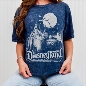 Pode incluir: Camiseta azul com um gr&aacute;fico branco do castelo da Disneylandia sob uma lua cheia e morcegos. O texto "Disneyland" e "I survived the happiest place on Earth" tamb&eacute;m est&atilde;o impressos na camiseta.