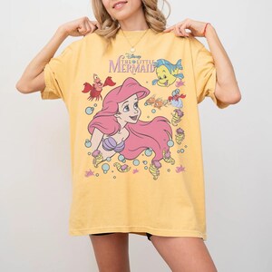 Pode incluir: Camiseta amarela mostarda com um gr&aacute;fico de Ariel de A Pequena Sereia da Disney. O design inclui Ariel com cabelo rosa, um suti&atilde; de concha roxa e v&aacute;rias criaturas marinhas. A camisa tem o logotipo do filme em roxo.