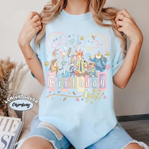 May include: Pale blue t-shirt with a vibrant graphic of cartoon characters, the words "Birthday Girl," and the name "Cali" with the number "3." The shirt is a Comfort Color Chambray.